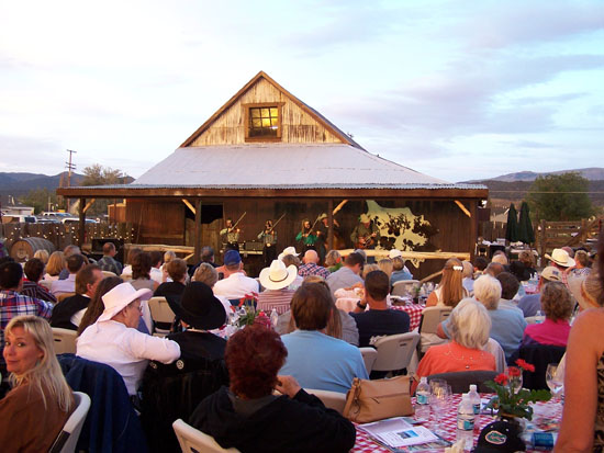 crowd of people at tables with red and white checked tablecloths watching musicians in front of a wooden building