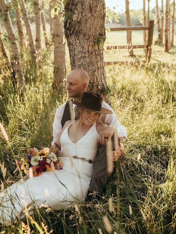 Bride and groom leaning against a tree. She is holding a floral bouquet and is waring a brown cowboy hat.