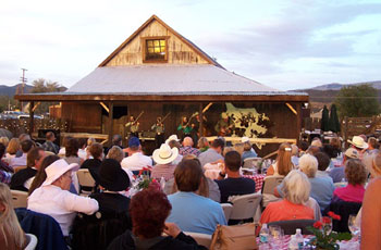 Large group of people sitting around tables at outdoor event in front of barn with musicians.